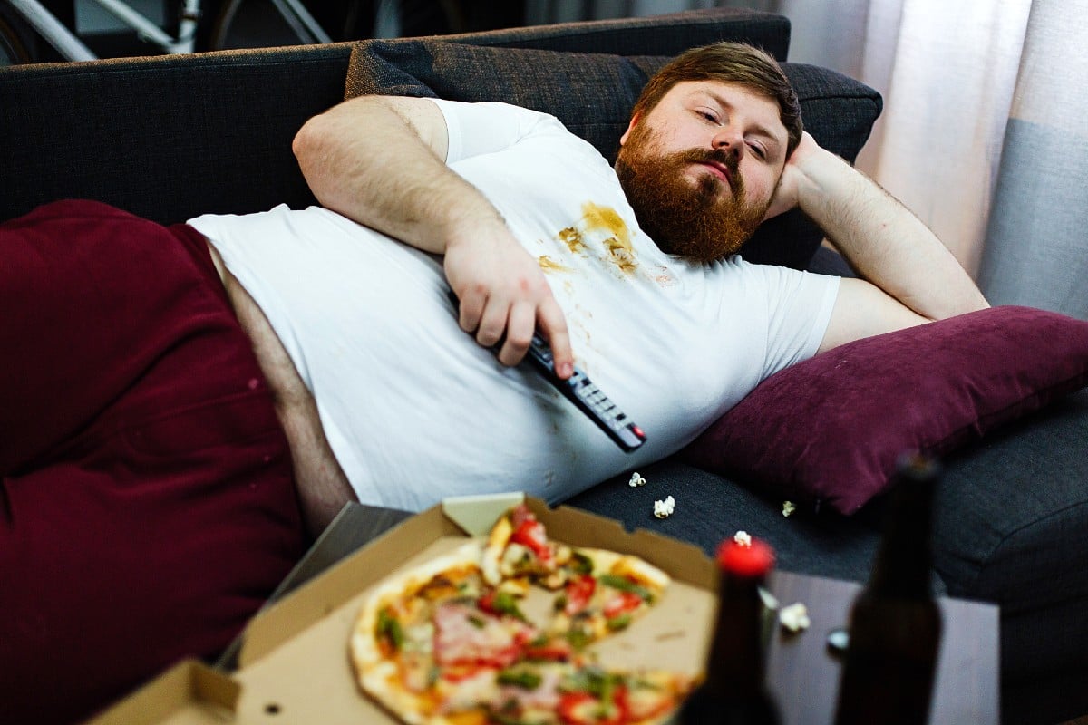 Un uomo grasso e pigro che mangia la pizza mentre è sdraiato sul divano a guardare la TV.